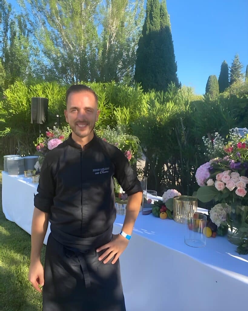 Barman souriant à un événement extérieur chic Un serveur souriant en chemise noire et tablier pose à côté d'une longue table blanche décorée de fleurs roses et jaunes, sous un ciel bleu.