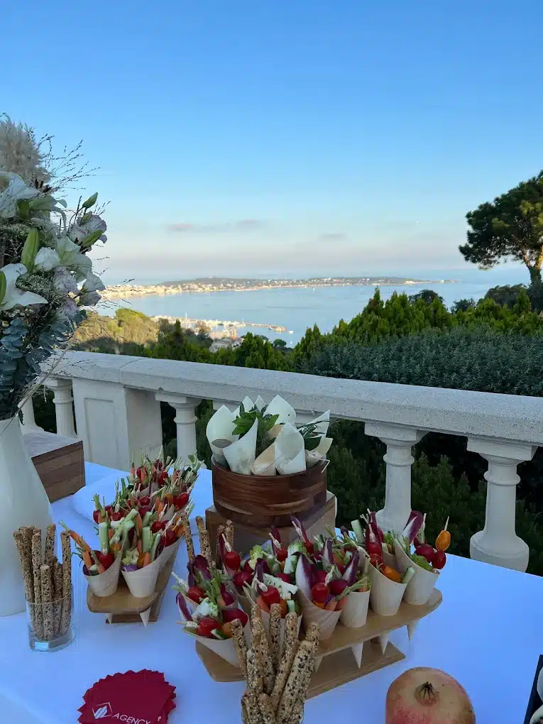 Buffet de légumes frais et gressins sur une table blanche, avec vue panoramique sur une baie et une ville côtière. Serviettes rouges "AGENCY".