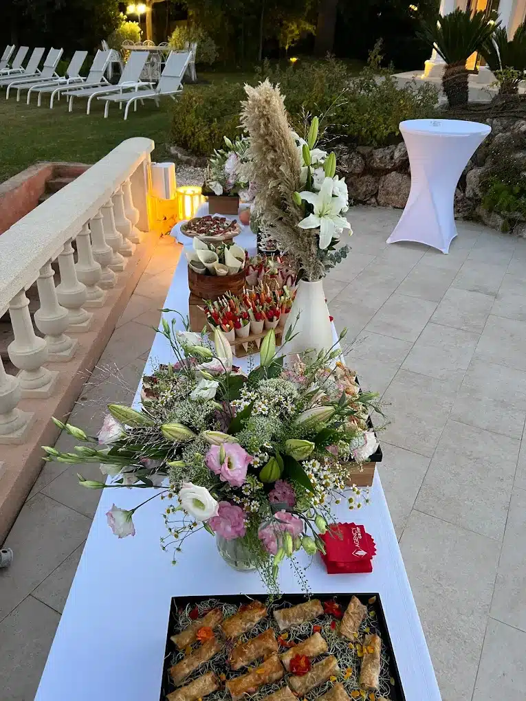 Buffet extérieur élégant au crépuscule avec de larges compositions florales blanches et roses, amuse-gueules et feuilletés, près de chaises longues.