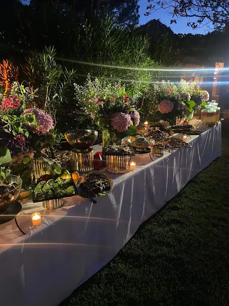 Buffet extérieur nocturne élégant avec fleurs, bougies et plats sur une longue table nappée de blanc, éclairé par des spots.