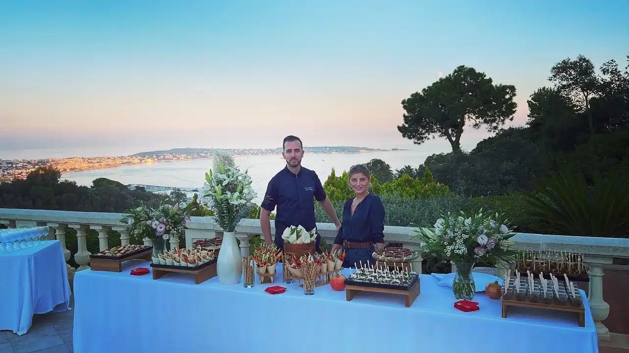 Deux traiteurs posent derrière un buffet de canapés élégants sur une terrasse avec vue panoramique sur la mer au coucher du soleil.