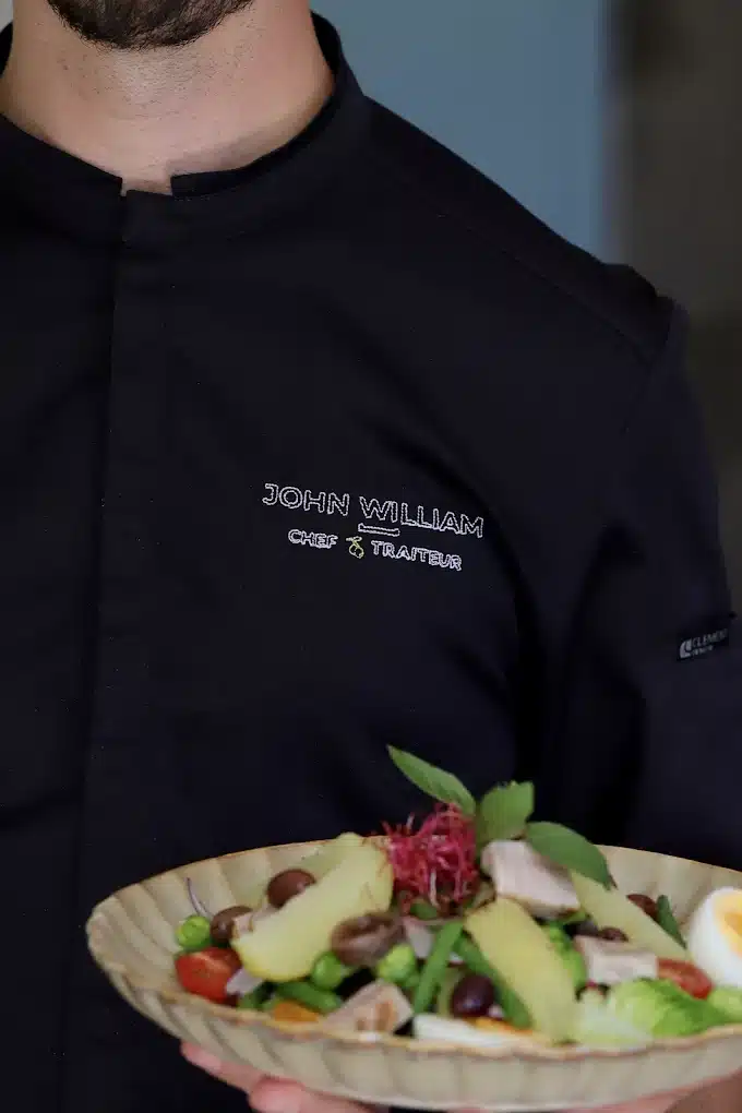 Chef en veste noire brodée "JOHN WILLIAM CHEF TRAITEUR" présentant une salade composée colorée, type salade niçoise, dans un plat texturé.
