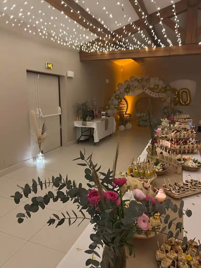 Ambiance de fête chaleureuse avec guirlandes lumineuses au plafond, buffet garni, et bouquet de roses roses. Décoration "Happy Birthday 50".