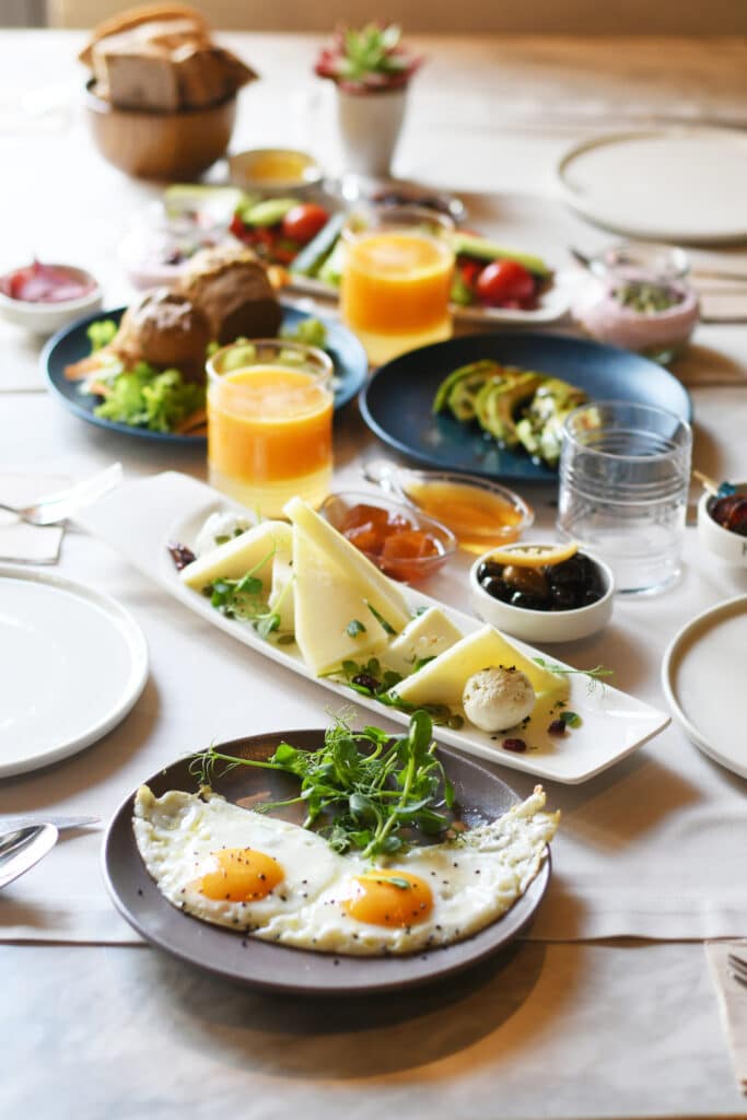 Petit-déjeuner complet : œufs, fromage et jus d'orange Petit-déjeuner copieux avec deux œufs au plat garnis de pousses, fromage, confiture, olives et jus d'orange sur une table claire.