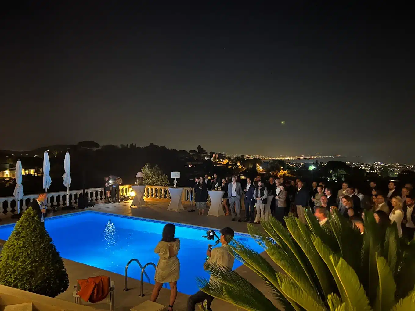 Soirée élégante au bord d'une piscine illuminée, avec invités en tenue de cocktail et vue nocturne panoramique sur une ville côtière éclairée.