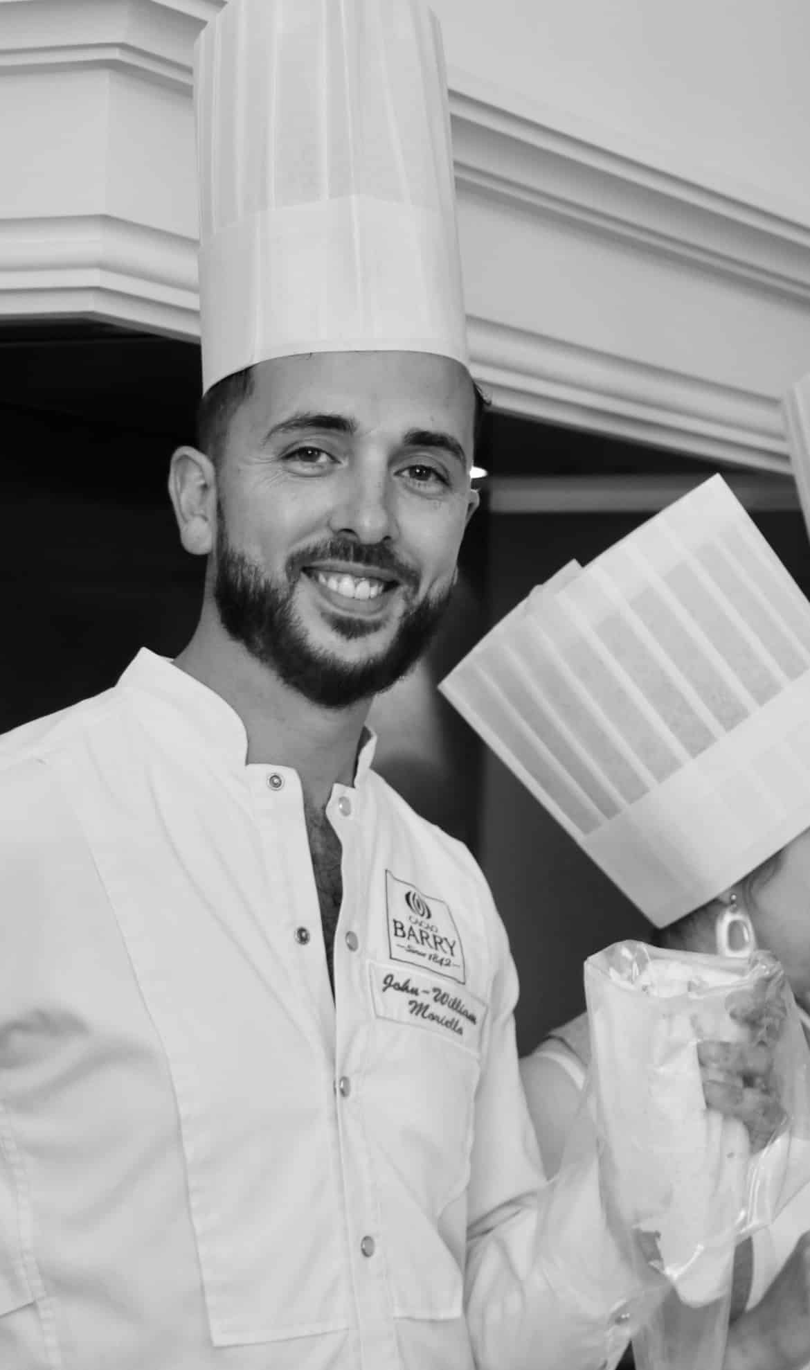 Chef John-William Moriella souriant en veste Cacao Barry avec toque, tenant une poche à douille.