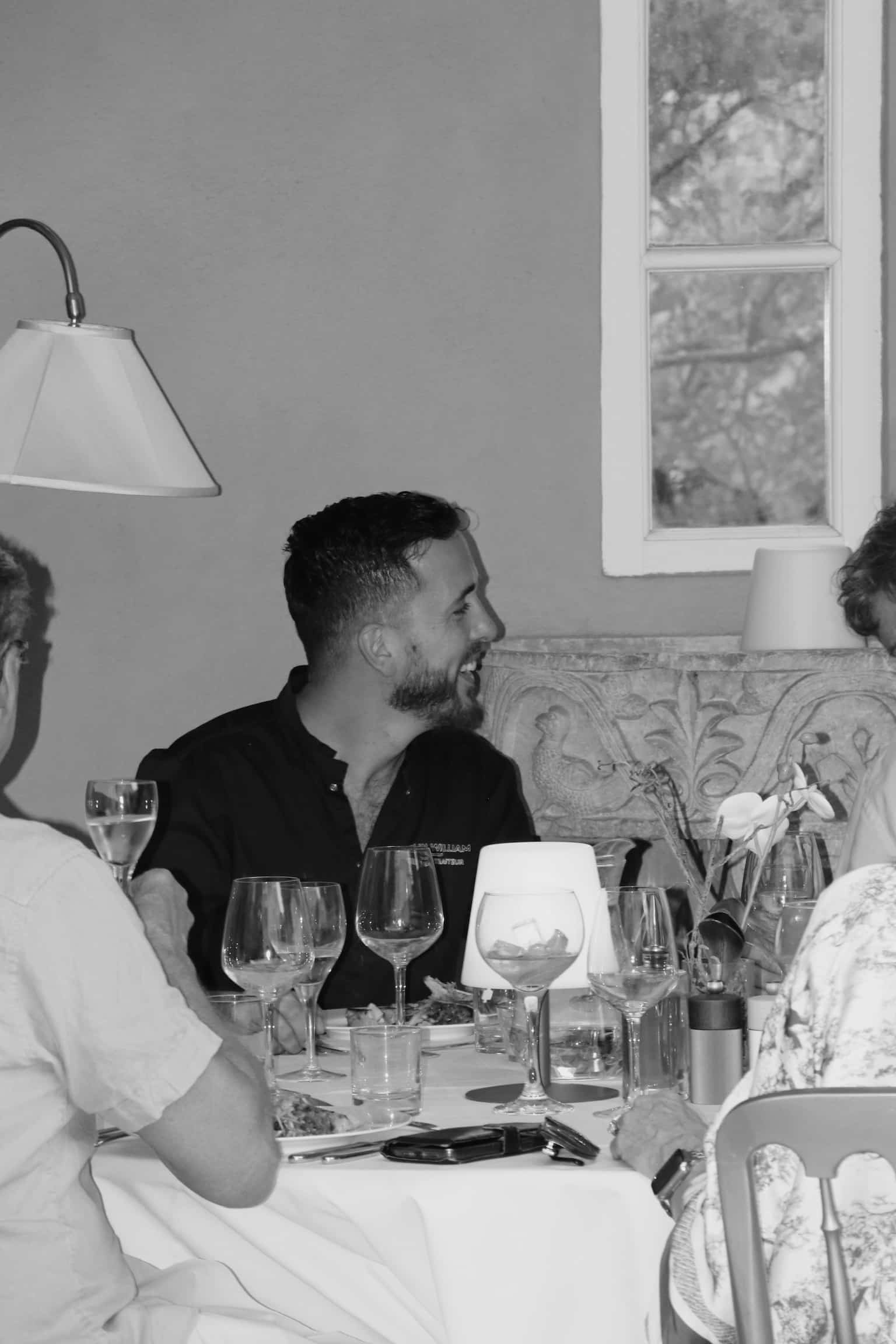Homme portant une chemise Jean William Traiteur souriant à table au restaurant. Scène conviviale en noir et blanc.