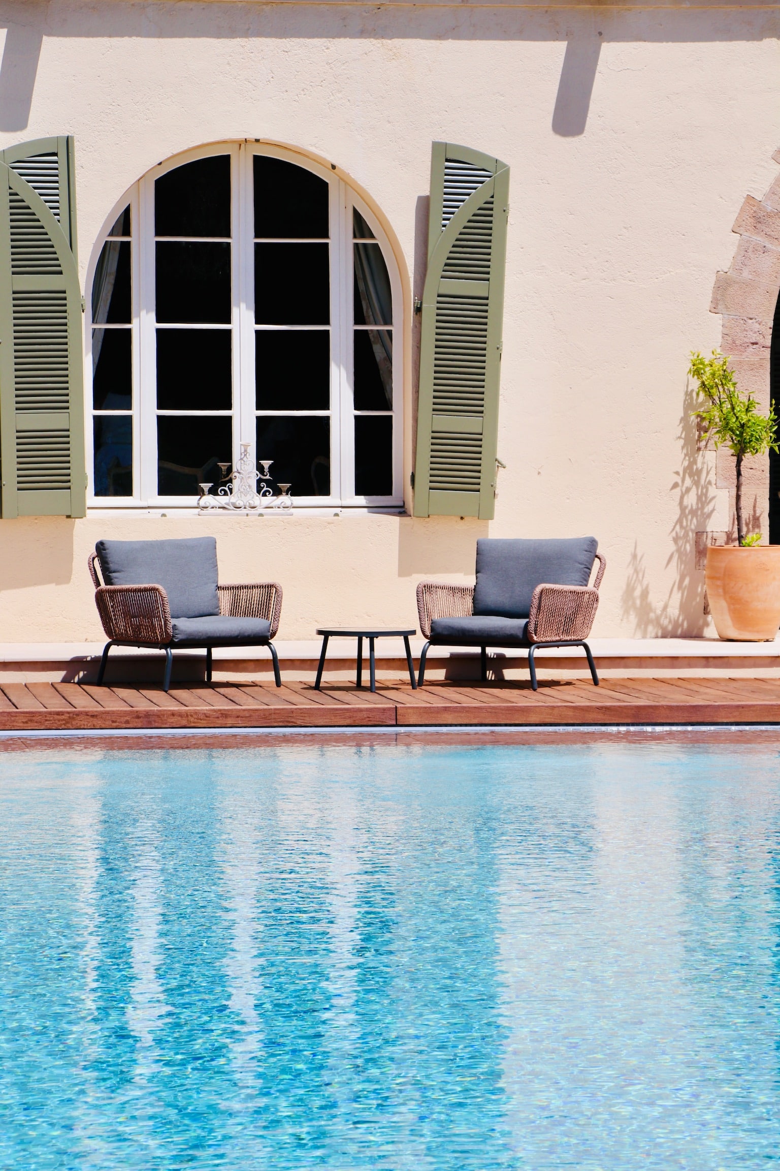 Fauteuils design et table basse sur terrasse en bois au bord d'une piscine ensoleillée devant une villa.