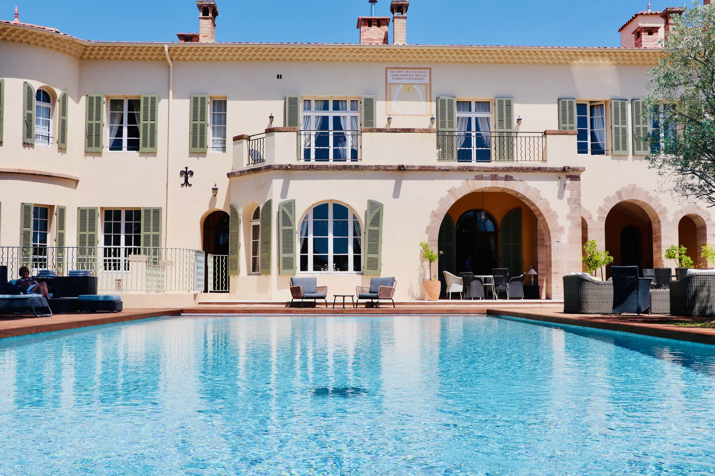 Piscine bleue devant une villa provençale aux volets verts et cadran solaire 'Gai lesert, beu ton souleu'.